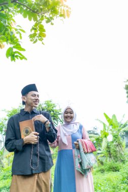 Bir çift genç Endonezyalı kadın ve erkek Müslüman kıyafetleri giyip Kuran 'a gidiyorlar. Kur 'an ve namaz araçlarını taşırlar..