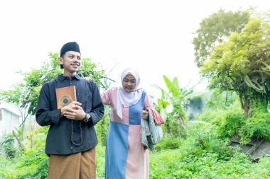 Bir çift genç Endonezyalı kadın ve erkek Müslüman kıyafetleri giyip Kuran 'a gidiyorlar. Kur 'an ve namaz araçlarını taşırlar..