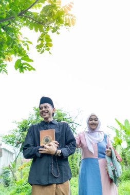 Bir çift genç Endonezyalı kadın ve erkek Müslüman kıyafetleri giyip Kuran 'a gidiyorlar. Kur 'an ve namaz araçlarını taşırlar..