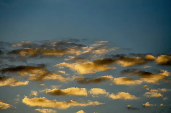 Derin mavi gökyüzü ve batan güneşin altın renklerini yakalayan beyaz bulutlarla inanılmaz bir akşam manzarası. Yumuşak alacakaranlık ışığı bulutlara sıcak ve altın bir ton verir, doğanın çarpıcı bir görüntüsünü yaratır..