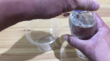 Asyalı bir adamın eli küçük bir şişeden ahşap bir masanın üzerindeki küçük plastik bir bardağa su döküyor.