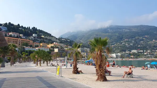 Haziran 2023 'te güneşli bir günde Menton' da plaj. Ön planda palmiye ağaçları, arka planda Fransız Alpleri görünür..