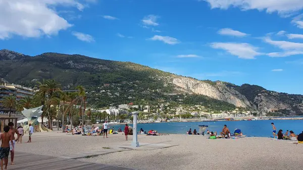 Akdeniz kıyısındaki Menton (Fransa) plajı, Ağustos 2023. Arkadaki kayalık dağlarda güneş parlıyor..