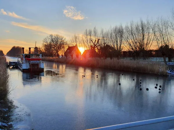 Flanders 'da (Belçika) kış günü gün batımında donmuş kanal. Bir grup kuş ve buharlı bir tekne, arka planda turuncu bir gökyüzüne karşı yel değirmenleri ve sıra sıra ağaçlar. Şubat 2021.