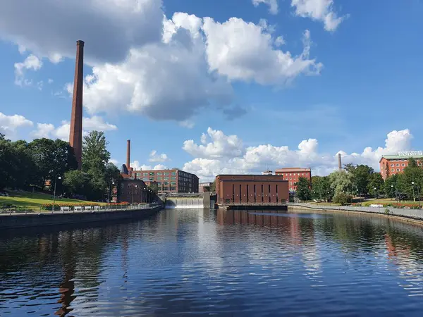 Tammerkoski, Finlandiya 'nın Tampere kentinde (Temmuz 2021) akıntıya kapıldı. Gökyüzünde beyaz bulutlar olan berrak mavi bir su. Uzun bacaları olan kırmızı tuğla fabrikaları. Nehrin kenarındaki çimen ve çiçek tesisleri.