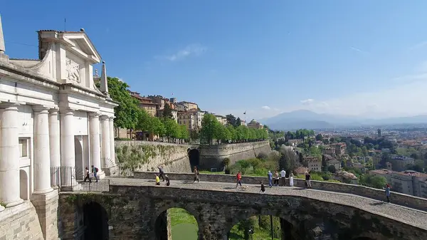 İtalya 'nın Bergamo kentini çevreleyen Venedik Duvarları (Citta Alta). Taştan bir köprüde beyaz bir şehir kapısına doğru yürüyen turistler. UNESCO Dünya Mirası sitesinin bir parçası. Bir bahar gününde şehrin kuş bakışı görüntüsü. Nisan 2024.