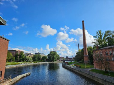 Tampere, Finlandiya (Ağustos 2024). Finlayson bölgesinden Tammerkoski nehri akıntıları görüldü. Temiz mavi suyun üzerinde kabarık beyaz bulutlar, bir tuğla baca ve yeşil bir park..