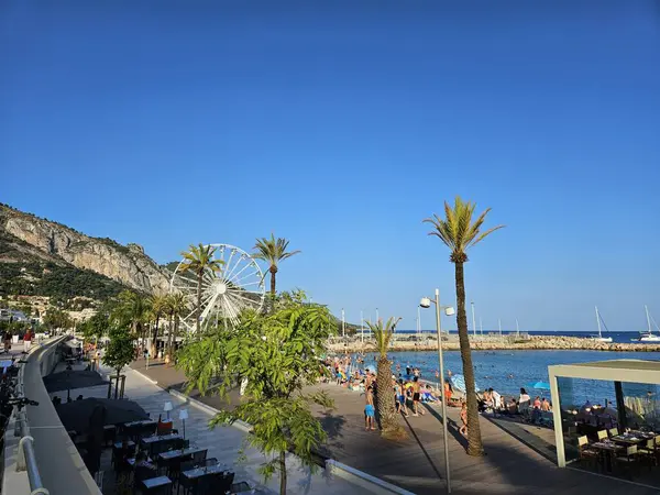 Menton, Fransa 'da sahil bulvarı (Ağustos 2024). Akdeniz kıyısında ahşap ve asfalt kaldırımlar, palmiye ağaçları. Dönme dolap ufukta.