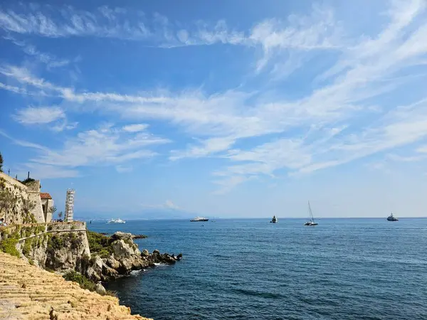 Antibes, Fransa (Ağustos 2024). Aydınlık bir yaz gününde Akdeniz manzarası. Denize bakan taş duvar ve kayalar, suda süzülen gemiler ve yelkenliler..