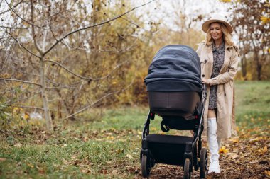 Genç anne, bebek arabasıyla ve yeni doğan çocuğuyla parkta yürüyor.