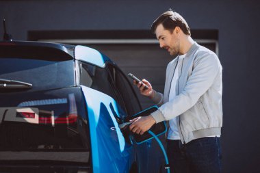 Genç adam evde elektrikli arabayı şarj ediyor ve cep telefonu kullanıyor.