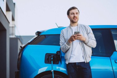 Genç adam evde elektrikli arabayı şarj ediyor ve cep telefonu kullanıyor.