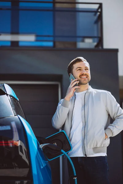 Młody Człowiek Ładuje Samochód Elektryczny Domu Używa Telefonu Komórkowego — Zdjęcie stockowe