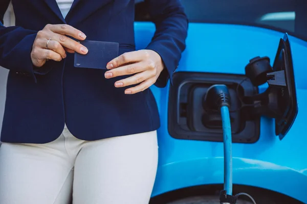Business woman standing by her electric car holding credit card