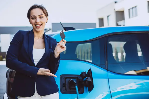 Business woman standing by her electric car holding credit card