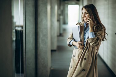 İş kadını elinde klasörler, telefon kullanarak ofiste yürüyor.