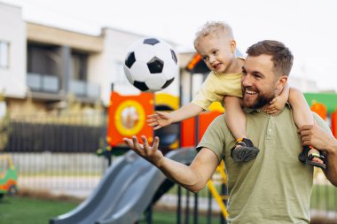 Baba oğul futbol topu tutuyorlar ve parkta eğleniyorlar.