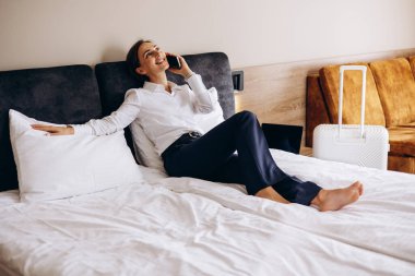 İş kadını bir otel odasında yatakta uzanıyor ve telefonla konuşuyor.