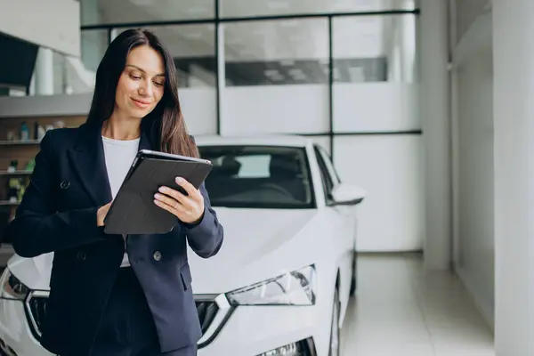 Sales woman with tablet in a car showroom standing by white car