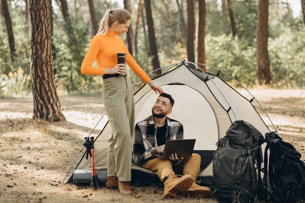 彼女のボーイフレンドがテントに座って森でコンピュータに取り組んでいる間 サーモスからお茶を飲む女性の旅行者 — ストック写真