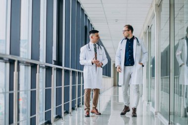 İki erkek doktor, klinikte yürüyorlar, beyaz laboratuvar önlüğü giyiyorlar.
