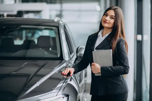 Young sales woman at carshowroom standing by the car