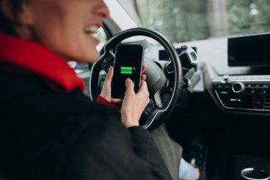 Bir kadın arabasını şarj ediyor ve telefonundaki çalara bakıyor.