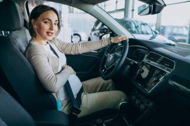 Genç hamile kadın araba galerisinde bir arabayı test ediyor.