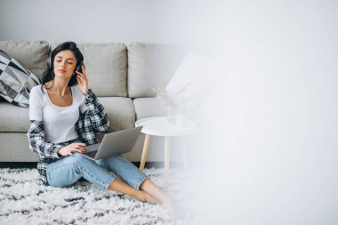 Genç ve güzel bir kadın evde oturmuş kulaklıkla dizüstü bilgisayarla çalışıyor.