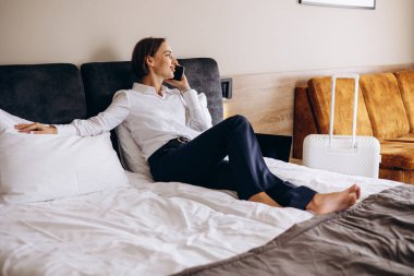 İş kadını bir otel odasında yatakta uzanıyor ve telefonla konuşuyor.