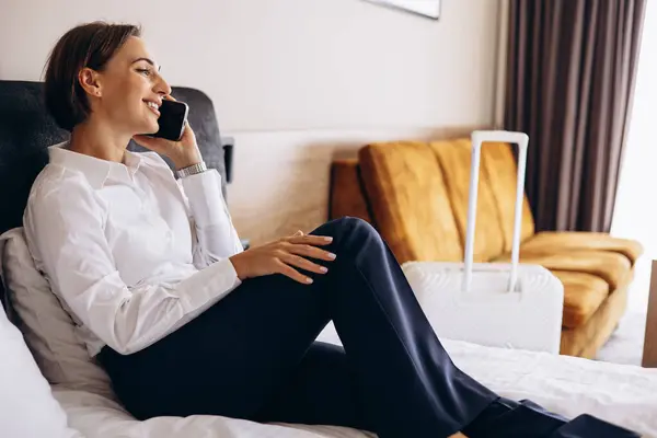 İş kadını bir otel odasında yatakta uzanıyor ve telefonla konuşuyor.