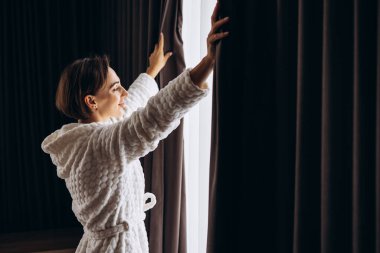 Otel odasında perdeleri açan bornozlu kadın.