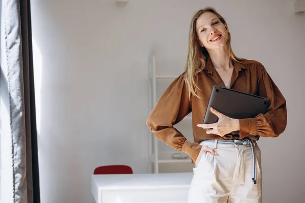 Business woman standing by the desk holding laptop at her home office