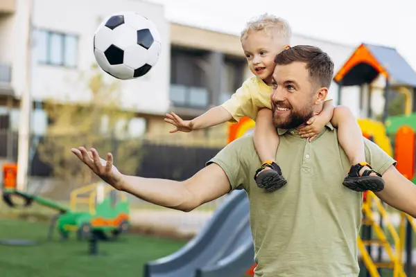Baba oğul futbol topu tutuyorlar ve parkta eğleniyorlar.
