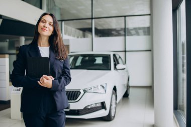 Tabletli bir kadın araba galerisinde beyaz arabanın yanında duruyor.