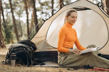 Genç bir kadın gezgin çadırının yanında kitap okuyor.