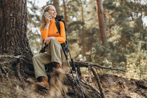 バッグ付きの女性旅行者とウォーキングスティック 木のそばに座って電話を使用 — ストック写真