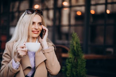 Kafenin dışında kahve içip telefonda konuşan yaşlı bir iş kadını.