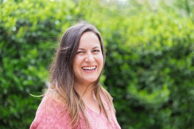 Outdoor portrait of a laughing 45 year old woman with windblown brown hair looking at the camera in the park on a sunny day. Confidence and tranquility of a mature woman. Happiness, lifestyle. clipart