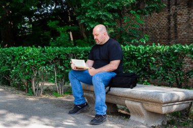 Güneşli bir öğleden sonra açık hava bankında oturmuş kitap okuyan 45-50 yaşlarında kel bir adamın portresi. Açık havada güneşli bir öğleden sonra kitap okumaya odaklanmış.
