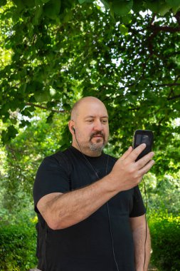 Kel, 45-50 yaşlarında, bıyıklı, keçi sakallı, siyah tişörtlü ve cep telefonuna kulaklıkla bakan bir adam.