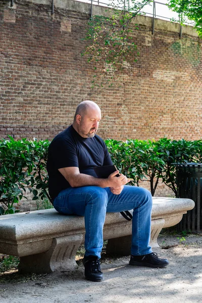 45-50 years old bald man in black T-shirt sitting on a park bench outdoors concentrating reading a book