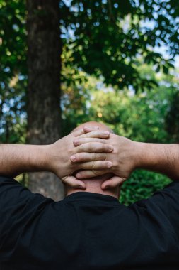 Elleri ensesinde olan, saçı az, saçı dökülmüş, olgun bir adamın arkasına bakın. Açık havada doğal ışık alan dikey fotoğrafçılık. Yansıma, gözlem ve dinlenme kavramı.