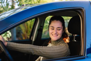 Güzel, genç bir kadın sürücü mutlu ve arabasının içinde gülümsüyor. Elleri direksiyonda ve geriye bakıyor. Yaşam tarzı ve sürüş kavramı