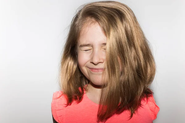 Güzel gülümseyen mutlu 8 yaşında, gözleri kapalı, somon rengi tişörtü ve beyaz arka planda yüzünün yarısını kaplayan sarı saçları olan bir kız çocuğu. Çocuklar ve çocukluk kavramı