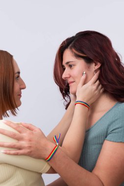 Öpüşmeden hemen önce, bileklerinde gökkuşağı bilezikleri ve güzel manikürleriyle birbirlerinin gözlerinin içine bakan genç, kızıl saçlı lezbiyen çiftin yakın çekim görüntüsü. Beyaz arka plan. LGTBI gurur ve haklar kavramı