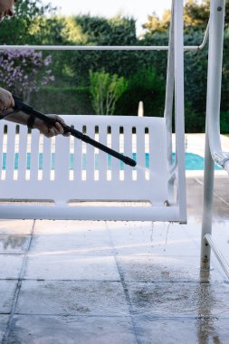 Özel bir evin havuzunda, bir yaz öğleden sonra, beyaz bir bahçe sallanan sandalyeyi basınç makinesiyle temizleyen bir adamın ellerinin yakın çekimi. Yaz konsepti ve ev işi
