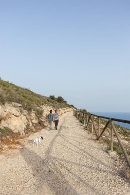 Bir oğlan ve bir kız arkalarında köpekleri, bir tarafta deniz ve gün batımında mavi gökyüzü ile dağların arasında geriye doğru yürüyorlar.