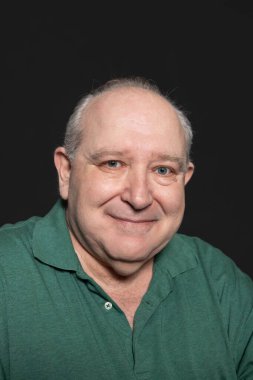 Yeşil polo tişört giymiş, gri saçlı ve mavi gözlü, rahat ve mutlu bir ifadeyle gülümseyen kameraya bakan seksiajiyen bir adamın portresi. Yaşlanma kavramı