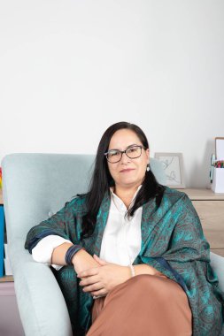 50 yaşında kadın psikoloji uzmanı, gözlüklü ve ipek eşarplı, gülümseyen ve rahat bir şekilde kameraya bakan, ofisindeki koltukta oturan. Kadın ve profesyonel kavramı